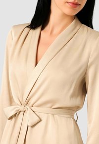 Vestito avvolgente beige con scollatura a V, maniche larghe e vita annodata. Tessuto liscio con una sottile lucentezza e dettagli minimali.