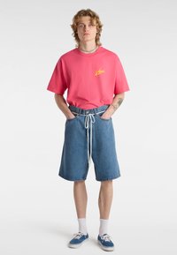 Różowy bawełniany t-shirt z żółtym logo, zestawiony z niebieskimi szortami dżinsowymi z troczkiem i podwiniętymi nogawkami. Stylizację dopełniają białe skarpetki i niebieskie sneakersy.