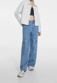 Veste courte gris clair avec fermeture à gros boutons, associée à un pantalon cargo en denim bleu ample avec de multiples poches et une coupe large.
