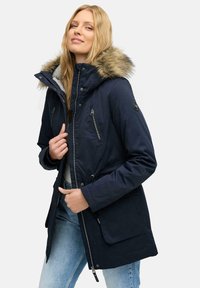 Navy parka jacket with faux fur trim, zippered pockets, drawstring waist, and soft lining. Worn over a light top and denim jeans.