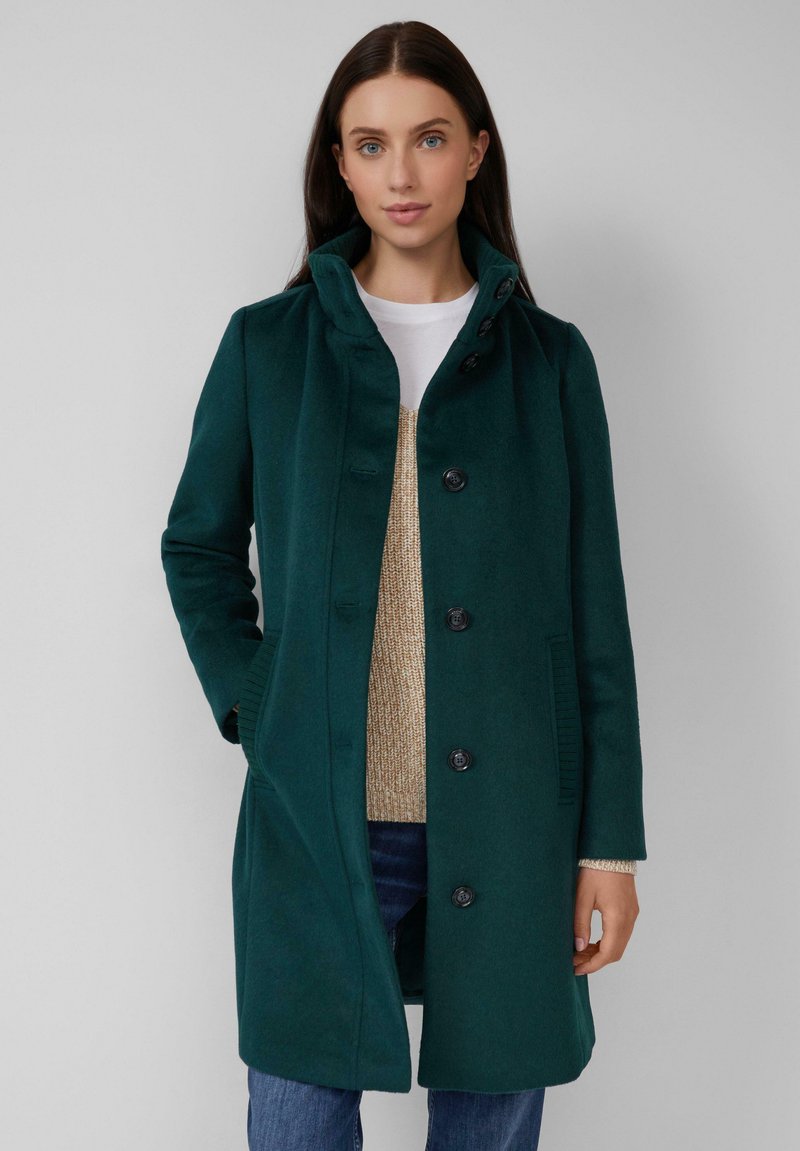 Dunkelgrünener Wollmantel mit hohem Kragen, knopfgeleitetem Vorderteil und seitlichen Taschen. Darunter wird ein beiger Strickpullover und blaue Jeans getragen.