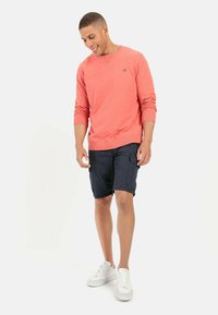 Koraalrood katoenen sweatshirt met lange mouwen en ronde hals, gecombineerd met donkere cargoshorts en witte sneakers, staande op een effen achtergrond.