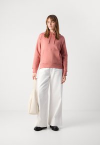 Giovane donna in felpa rosa e pantaloni larghi bianchi, che tiene una borsa in tela beige, in piedi contro uno sfondo bianco uniforme.