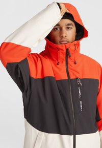 Chaqueta O'Neill colorida que presenta una mezcla de paneles naranjas, negros y blancos, con cierre frontal con cremallera, capucha ajustable y tejido texturizado.