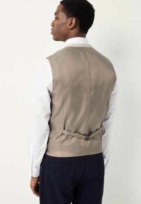 Gilet beige à la texture lisse, avec une sangle ajustable à l'arrière, porté par-dessus une chemise blanche à manches longues, associé à un pantalon sombre.