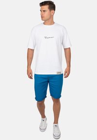 Witte t-shirt met korte mouwen en een zwarte logoprint, gecombineerd met blauwe knielange shorts met opgerolde pijpen en contrasterende accenten, gedragen met witte schoenen.