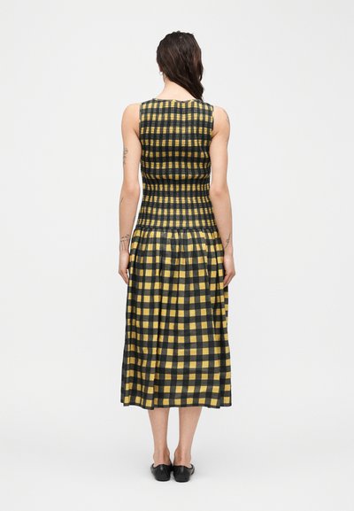 Γυναίκα που στέκεται με την πλάτη γυρισμένη, φορώντας μίντι φόρεμα χωρίς μανίκια σε κίτρινο και μαύρο καρό με σούρες στη μέση και μαύρα επίπεδα παπούτσια.