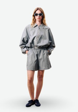 Femme portant une veste grise surdimensionnée à fermeture éclair, un short taille haute assorti, des lunettes de soleil bleues et des baskets bleu marine, debout les mains dans les poches.