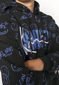 Černá mikina s kapucí pokrytá modrými usmívajícími se obličeji a nápisem "Karl Kani", s velkým modro-bílým designem "SMILE" na přední straně, nošena se zkříženýma rukama.