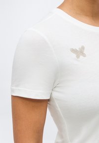 Λευκό πουκάμισο με κοντά μανίκια που φοριέται στο πάνω μέρος του σώματος, με μια μικρή καρφίτσα σε σχήμα πεταλούδας από συστάδες μαργαριταριών καρφωμένη κοντά στην κλείδα.