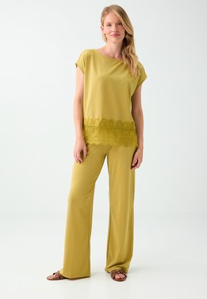 Femme aux longs cheveux blonds portant un pantalon large vert olive et un haut assorti avec un détail en dentelle, debout devant un fond blanc.