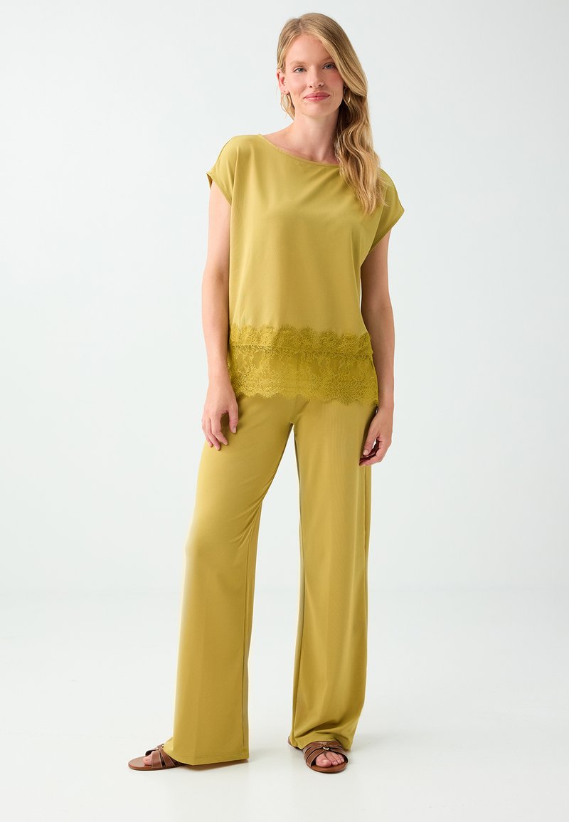 Femme aux longs cheveux blonds portant un pantalon large vert olive et un haut assorti avec un détail en dentelle, debout devant un fond blanc.