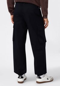 Schwarze Cargo-Hosen mit Seitentaschen, im entspannten Schnitt und mit elastischem Bund. Kombiniert mit weißen Turnschuhen.