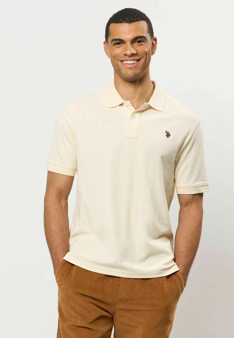 Homme souriant portant un polo beige clair à manches courtes avec un petit logo et un pantalon en velours côtelé marron, mains dans les poches.