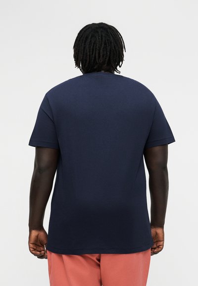 Person mit dunkler Haut und Dreadlocks, die von hinten zu sehen ist, trägt ein marineblaues Kurzarmhemd und korallenfarbene Hosen.