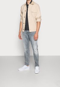 Beige jacka med två framfickor, svart polotröja, ljusblå jeans och vita sneakers. Slät tyg och avslappnad passform.