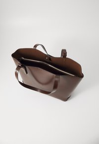 Sac fourre-tout en cuir brun avec deux poignées, ouverture sur le dessus et une poche intérieure zippée ; texture lisse et forme structurée.