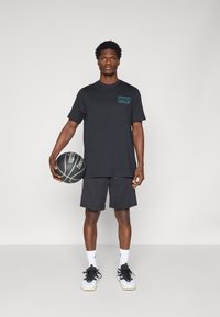 Zwart T-shirt met korte mouwen en een teal grafiek op de borst, gecombineerd met zwarte shorts en geaccentueerd met witte sokken en schoenen. Houdt een basketbal vast.