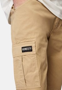 Cargo shorts i khaki-stof har en sidelomme med en sort etiket, der står "ÆRLIGHED ER REGELEN," hvilket fremhæver det praktiske design og syningerne.