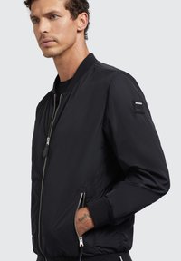 Jachetă bomber neagră cu fermoar frontal, guler și manșete ribbed, dotată cu buzunare laterale și un patch cu logo pe mâneca stângă. Textură netedă.