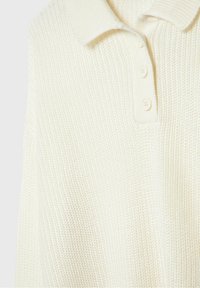 Gebreide trui in roomkleur met een geribbelde textuur, voorzien van een kraag en een drieknoopslijst. De stof oogt zacht en dik.