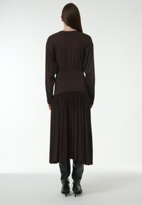 Dunkelbraunes, langärmliges Kleid mit tailliertem Schnitt und vollem, plissiertem Rock. Getragen mit kniehohen schwarzen Stiefeln. Glatte Textur, minimalistische Details.