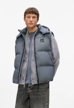 Ung mand iført grå polstret vest over en lagdelt grå hættetrøje og t-shirt, stående mod en enkel hvid baggrund.