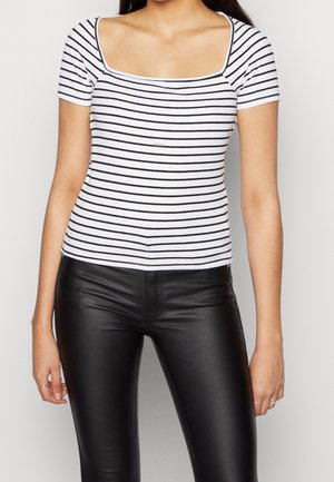 Femme portant un haut blanc à manches courtes avec des rayures horizontales noires et un pantalon en cuir noir ajusté sur un fond uni.
