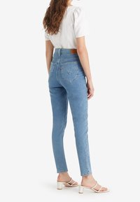 Mujer vestida con jeans ajustados de tiro alto en azul claro, blusa blanca de manga corta y sandalias de tacón blancas, de pie frente a un fondo blanco.