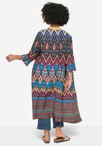 Värikäs kimono-tyylinen päällyste, jossa on geometrisia kuvioita punaisissa, sinisissä ja keltaisissa sävyissä, leviäthihnoilla ja avoimella edessä.
