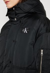 Chaqueta de plumas negra con un alto collar, textura brillante, botones de presión en la parte delantera y un bolsillo con cremallera lateral que presenta un parche de logo en el pecho.