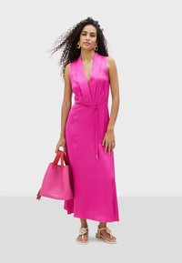 Vestido rosa sin mangas con escote en V, cintura ceñida y falda fluida. Acompañado de un bolso rosa con asas rojas y sandalias beige.