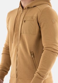 Felpa con zip color sabbia, realizzata in tessuto morbido, dotata di tasca frontale con zip, tasca laterale con zip e polsini a costine. Design semplice e minimalista.