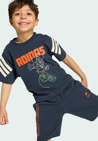 T-shirt di cotone blu navy con righe bianche, presenta una grafica di Topolino e "ADIDAS" in arancione. Pantaloni corti coordinati con il marchio "WALT DISNEY".