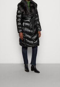 Abrigo de plumas negro con cuello de pelo sintético, cintura ceñida y textura brillante. Presenta un diseño acolchado y se combina con vaqueros oscuros y botines.