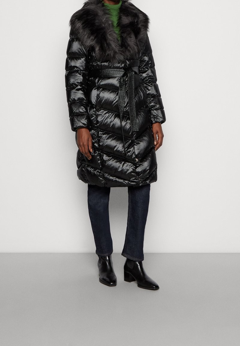 Abrigo de plumas negro con cuello de pelo sintético, cintura ceñida y textura brillante. Presenta un diseño acolchado y se combina con vaqueros oscuros y botines.