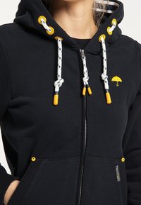 Felpa nera con cappuccio realizzata in tessuto morbido, con dettagli gialli sui lacci e sul logo, chiusura con zip e tasche frontali.
