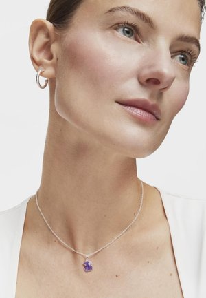 Collar de plata con un colgante de gema púrpura en forma de flor, que presenta una fina cadena y una vista en primer plano de la persona que lo lleva puesto.
