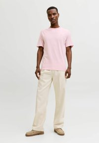 Hellrosa Baumwoll-T-Shirt mit Rundhalsausschnitt, kurzen Ärmeln und einem dezenten Logodetail. Kombiniert mit cremefarbenen Hosen und beigen Schuhen.