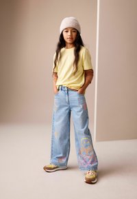 Lysegul T-shirt med korte ærmer, lyseblå vidde bukser med farverige smilende ansigter og hjerte-designs, strikket beige hue, flerfarvede sneakers.