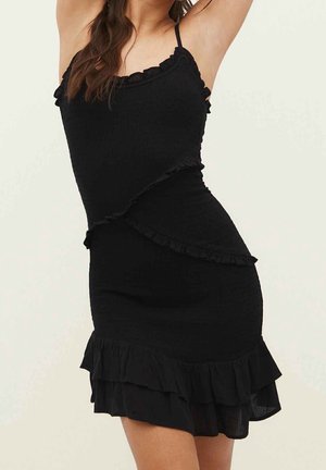 Kobieta w dopasowanej czarnej sukience bez rękawów z falbankami na krawędziach, pozująca z uniesionymi ramionami na jednolitym jasnym tle.