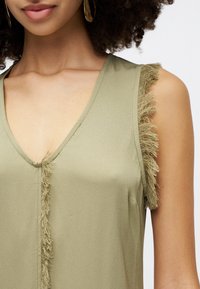 Oliwkowa bezrękawnik z głębokim dekoltem w kształcie litery V, z frędzlami wzdłuż otworów na ramiona, co dodaje tekstury i wizualnego zainteresowania.