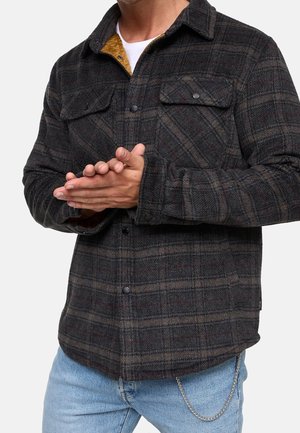 Mand iført mørk ternet skjorte med knapper, lyseblå jeans og sølvkæde, der holder hænderne foldet ved taljen.