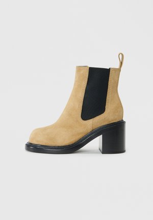 Bottine en suède beige avec un panneau latéral élastique noir, bout rond et talon épais noir, dotée d'une tirette à l'arrière.