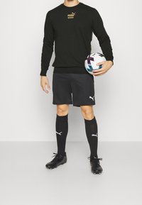 Felpa nera con logo dorato, pantaloni corti neri, calze nere e scarpini da calcio neri; tiene in mano un pallone da calcio multicolore.
