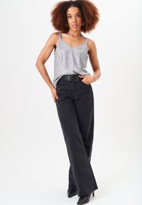 Young woman with curly hair wearing a silver sequin tank top, black wide-leg pants, black belt, and black pointed boots, posing confidently.