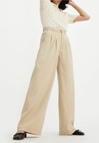 Beige vida byxor med veck, tillverkade av ett mjukt tyg, tillsammans med en top i spetsstickning och svarta slip-on skor.