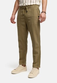 Pantalones jogger verde oliva hechos de tejido texturizado con una cintura ajustable por cordón, con bolsillos laterales y puños ajustados, llevados con zapatillas ligeras.