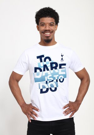 Uśmiechnięty mężczyzna z kręconymi włosami, ubrany w biały T-shirt Tottenham Hotspur z napisem "TO DARE IS TO DO", z rękami na biodrach, jasne tło.