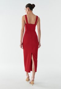 Rød tætsiddende midi-kjole med brede stropper, lynlås i ryggen og en høj slids til låret. Glat stof og struktureret design for et skræddersyet udseende.